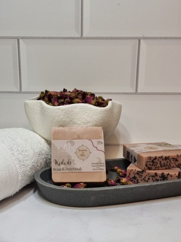 A tile background and stone bench with a grey oval dish. Three bars of pink homemade soap are sitting on the dish, with small red rose buds to decorate. Also in the background are a small white towel and a fluted white bowl full of small red rose buds