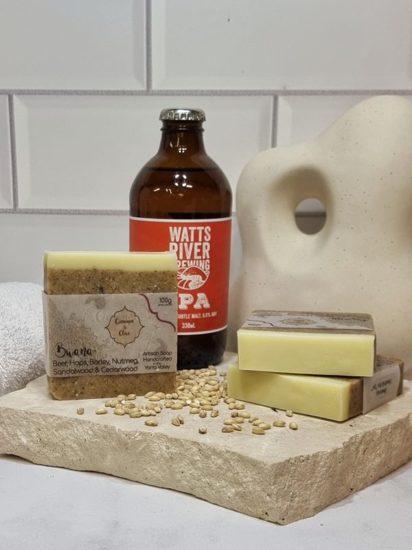 A tile background and stone bench with a piece of terrazzo. Three bars of pale tan and white layered homemade soap are sitting on the terrazzo, with barley grains and a bottle of Watts River IPA. Also in the background are a small white towel and a curvy decorative statue