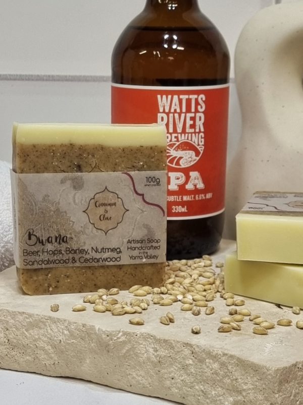 A tile background and stone bench with a piece of terrazzo. Three bars of pale tan and white layered homemade soap are sitting on the terrazzo, with barley grains and a bottle of Watts River IPA. Also in the background are a small white towel and a curvy decorative statue