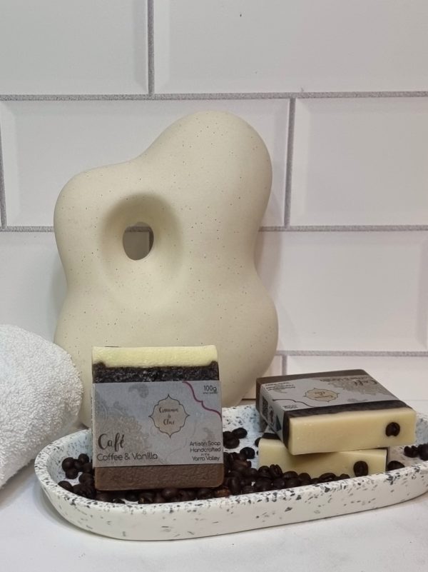 A tile background and stone bench with an oval terrazo dish. Three bars of cream and brown layered homemade soap are sitting on the dish, with coffee beans to decorate. Also in the background are a small white towel and a curvy decorative statue