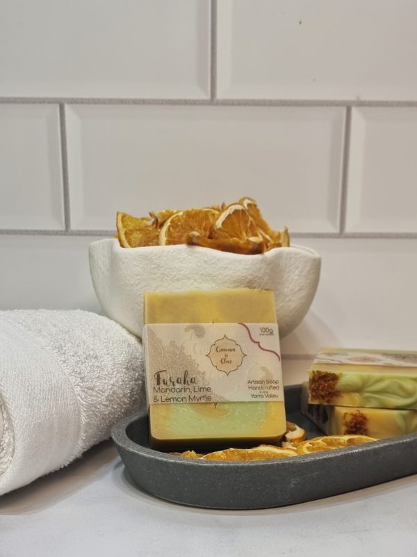 A tile background and stone bench with a grey oval dish. Three bars of orange, yellow and green swirled homemade soap are sitting on the dish, with dried orange slices to decorate. Also in the background are a small white towel and a fluted white bowl full of dried orange slices