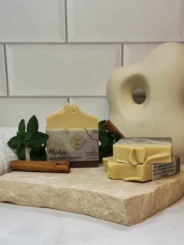 A tile background and stone bench with a piece of terrazzo. Three bars of cream, green and brown layered homemade soap are sitting on the terrazzo, with cinnamon sticks and fresh mint sprigs to decorate. Also in the background are a small white towel and a curvy decorative statue