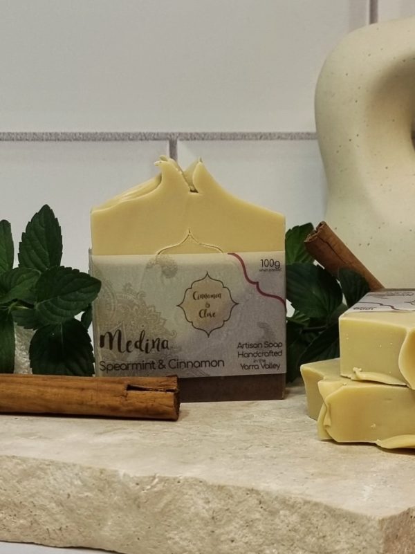 A tile background and a piece of terrazzo. Three bars of cream, green and brown layered homemade soap are sitting on the terrazzo, with cinnamon sticks and fresh mint sprigs to decorate. Also in the background are a small white towel and a curvy decorative statue