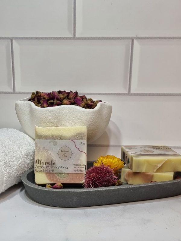 A tile background and stone bench with a grey oval dish. Three bars of cream, green, pink and purple swirled homemade soap are sitting on the dish, with dried orange and purple flowers to decorate. Also in the background are a small white towel and a fluted white bowl full of small red rose buds