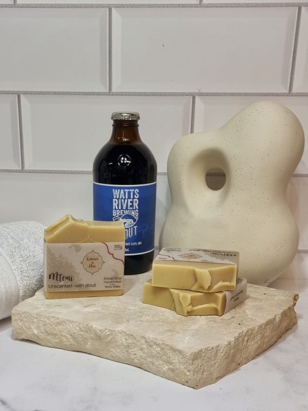 A tile background and stone bench with a piece of terrazzo. Three bars of pale tan coloured homemade soap are sitting on the terrazzo, with a bottle of Watts River Stout. Also in the background are a small white towel and a curvy decorative statue