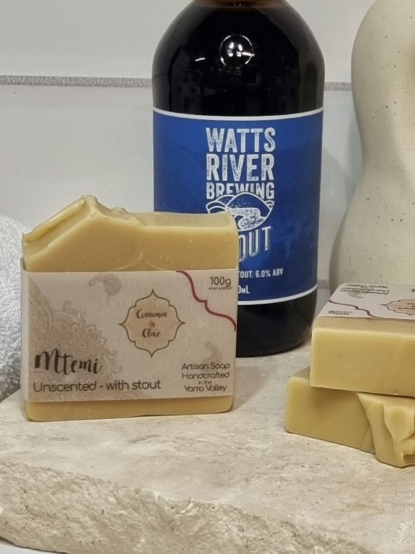 A tile background with a piece of terrazzo. Three bars of pale tan coloured homemade soap are sitting on the terrazzo, with a bottle of Watts River Stout. Also in the background are a small white towel and a curvy decorative statue
