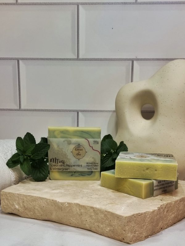 A tile background and a piece of terrazzo. Three bars of blue and creamy swirled homemade soap are sitting on the terrazzo, with fresh mint sprigs to decorate. Also in the background is and a curvy decorative statue