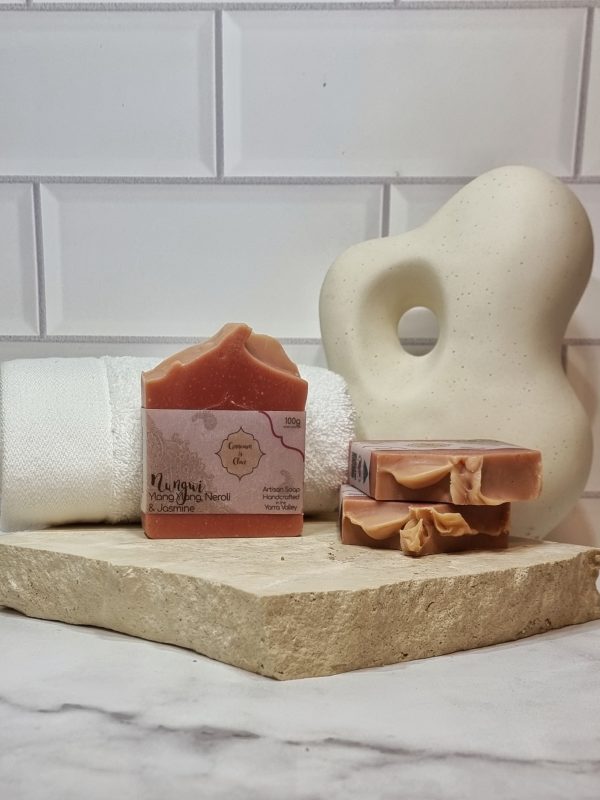 A tile background and stone bench with a piece of terrazzo. Three bars of bright orangey pink homemade soap are sitting on the terrazzo. Also in the background are a small white towel and a curvy decorative statue