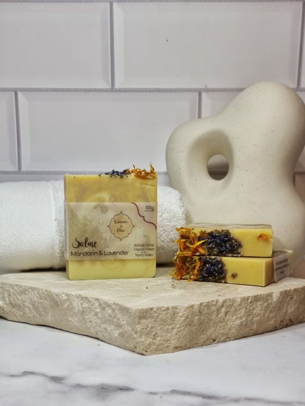 A tile background and stone bench with a piece of terrazzo. Three bars of cream, orange and purple swirled homemade soap topped with calendula petals and lavender buds are sitting on the terrazzo. Also in the background are a small white towel and a curvy decorative statue