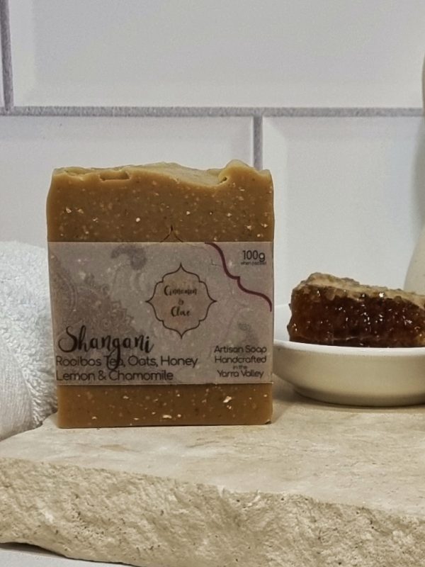 A tile background and a piece of terrazzo. One bar of honey coloured homemade soap is sitting on the terrazo, with a small white dish of honey on the comb to decorate. Also in the background is a small white towel