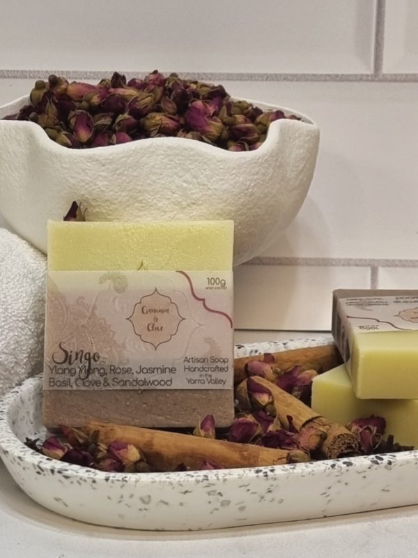 A tile background and stone bench with a white terrazo oval dish. Three bars of pink, brown and white layered homemade soap with a rosebud on top are sitting on the dish, with cinnamon sticks, clove buds and rose buds to decorate. Also in the background are a small white towel and a fluted white bowl full of small red rose buds