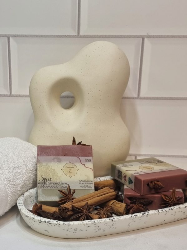 A tile background and stone bench with an oval terrazo dish. Three bars of red, white and brown layered homemade soap are sitting on the dish, with star anise, cinnamon sticks and clove buds to decorate. Also in the background are a small white towel and a curvy decorative statue