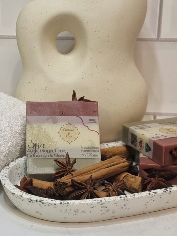 A tile background and stone bench with an oval terrazo dish. Three bars of red, white and brown layered homemade soap are sitting on the dish, with star anise, cinnamon sticks and clove buds to decorate. Also in the background are a small white towel and a curvy decorative statue