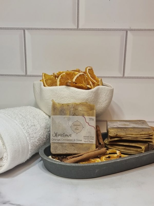 A tile background and stone bench with a grey oval dish. Three bars of orange and brown swirled homemade soap are sitting on the dish, with dried orange slices, cinnamon sticks and clove buds to decorate. Also in the background are a small white towel and a fluted white bowl full of dried orange slices