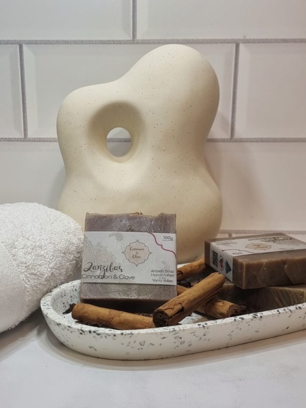 A tile background and stone bench with an oval terrazo dish. Three bars of brown swirled homemade soap are sitting on the dish, with cinnamon sticks and clove buds to decorate. Also in the background are a small white towel and a curvy decorative statue