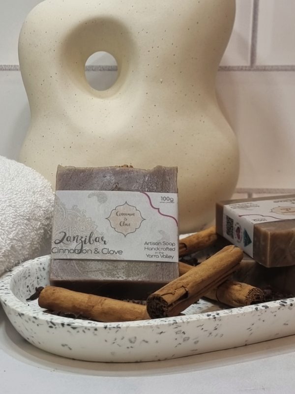 A tile background and stone bench with an oval terrazo dish. Three bars of brown swirled homemade soap are sitting on the dish, with cinnamon sticks and clove buds to decorate. Also in the background are a small white towel and a curvy decorative statue