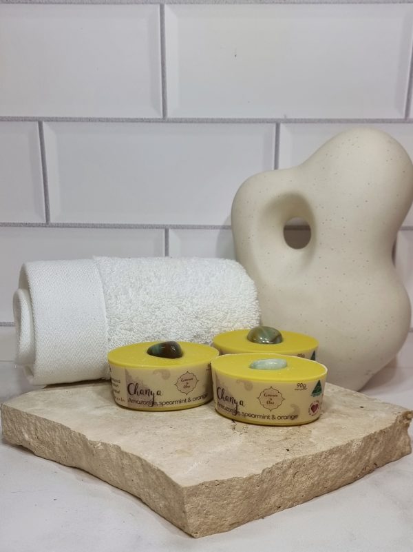 A tile background with a stone benchtop and a piece of terrazzo. Three bars of yellow coloured oval shaped soaps with a green crystal in top are sitting on the terrazzo. Also in the background are a small white towel and a curvy decorative statue