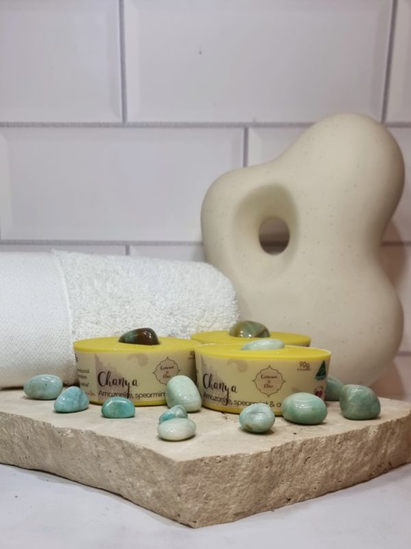 A tile background with a piece of terrazzo. Three bars of yellow coloured oval shaped soaps with a green crystal in top are sitting on the terrazzo. Some green crystals are scattered around the soaps. Also in the background are a small white towel and a curvy decorative statue