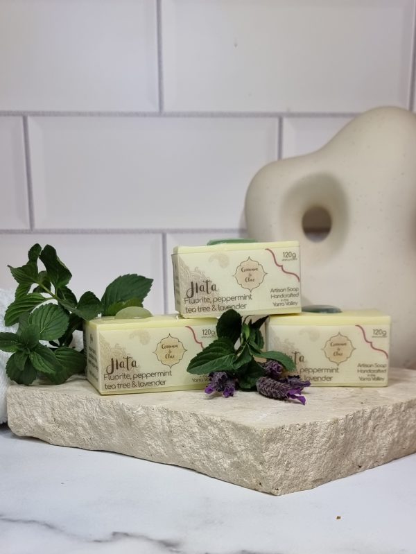 A tile background with a piece of terrazzo. Three bars of white square soaps with a green crystal in top are sitting on the terrazzo with fresh sprigs of lavender and mint. Also in the background are a small white towel and a curvy decorative statue
