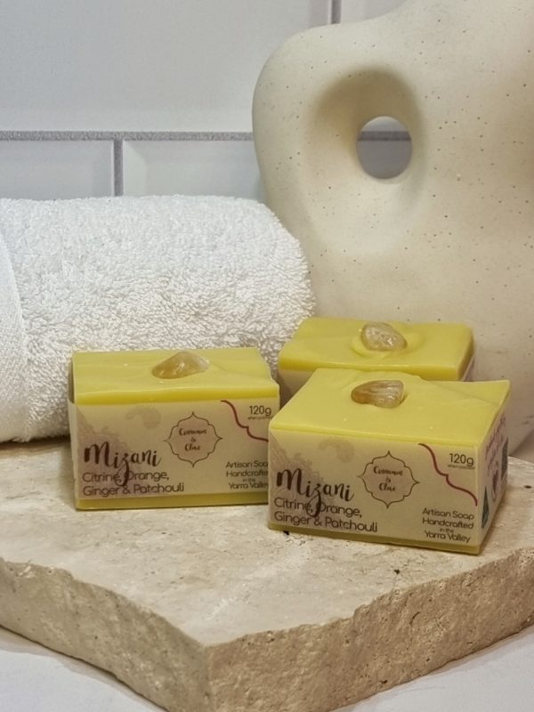 A tile background with a stone benchtop and a piece of terrazzo. Three bars of yellow coloured square soaps with a yellow crystal in top are sitting on the terrazzo. Also in the background are a small white towel and a curvy decorative statue