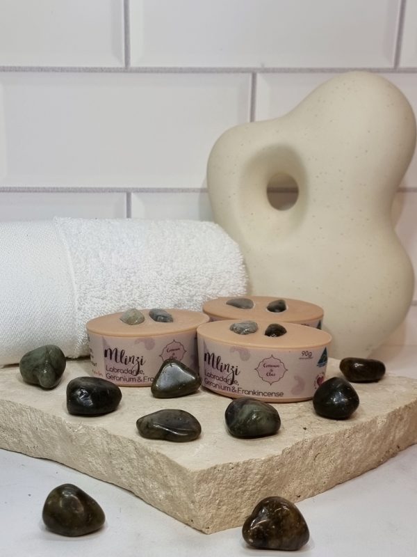 A tile background with a piece of terrazzo. Three bars of pink coloured oval shaped soaps with a dark grey crystal in top are sitting on the terrazzo with more dark crystals scattered around them. Also in the background are a small white towel and a curvy decorative statue