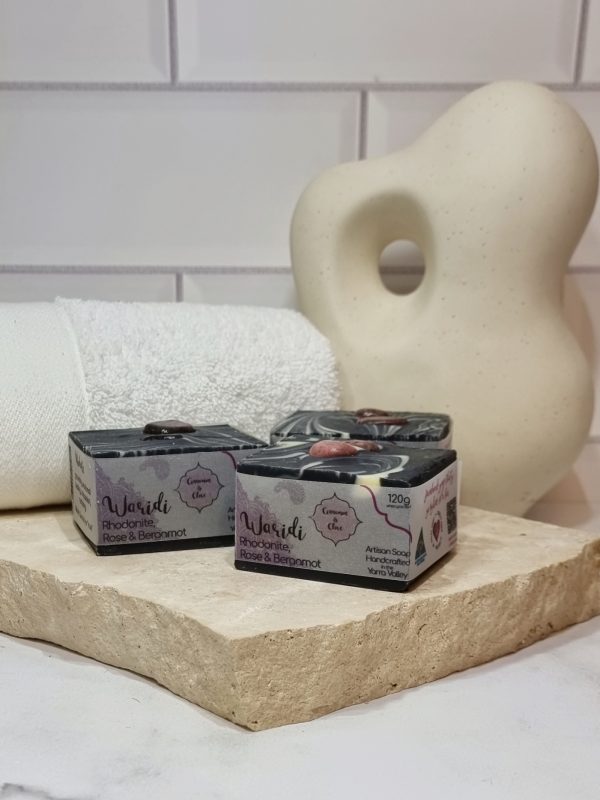 A tile background with a stone benchtop and a piece of terrazzo. Three bars of black square soaps with a white swirl and two pink crystal in the top of each are sitting on the terrazzo. Also in the background are a small white towel and a curvy decorative statue