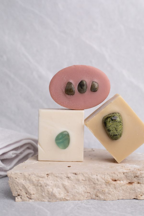 A stone backdrop with a piece of terrazzo on the benchtop. On the terrazzo are three crystal soaps - a white square soap with a green crystal, and a yellow square soap with a yellow and black speckled crystal, then on top of these two is a pink oval soap with three small dark grey crystals on top. In the background is a small white towel