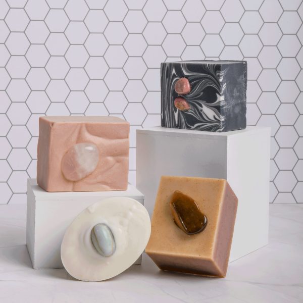 A tiled background and white benchtop with two small white boxes on top. On the boxes is a pink square soap and a black and white square soap, each with pink crystals in the top. In front is a white oval soap with green crystal and brown square soap with a brown crystal in the top