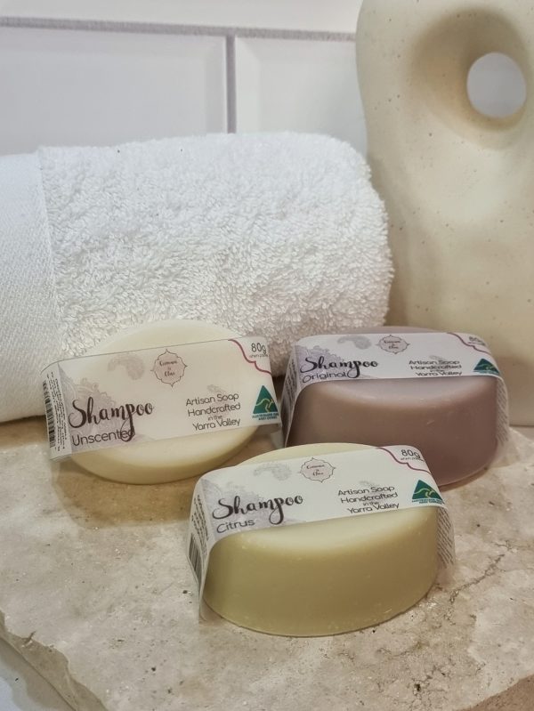 A tile background and a piece of terrazzo. Three bars of oval shaped shampoo bars are sitting on the terrazzo - one is white, one is purple and one is pale green. Also in the background are a small white towel and a curvy decorative statue