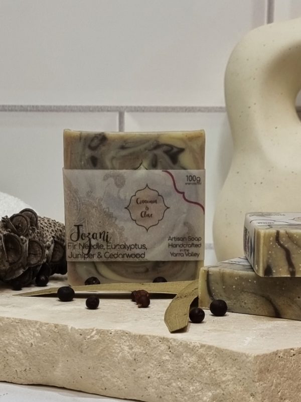 A tile background and a piece of terrazzo. Three bars of a camouflage design homemade soap are sitting on the terrazo, with juniper berries, seed pods and eucalyptus leaves to decorate. Also in the background are a small white towel and a curvy decorative statue