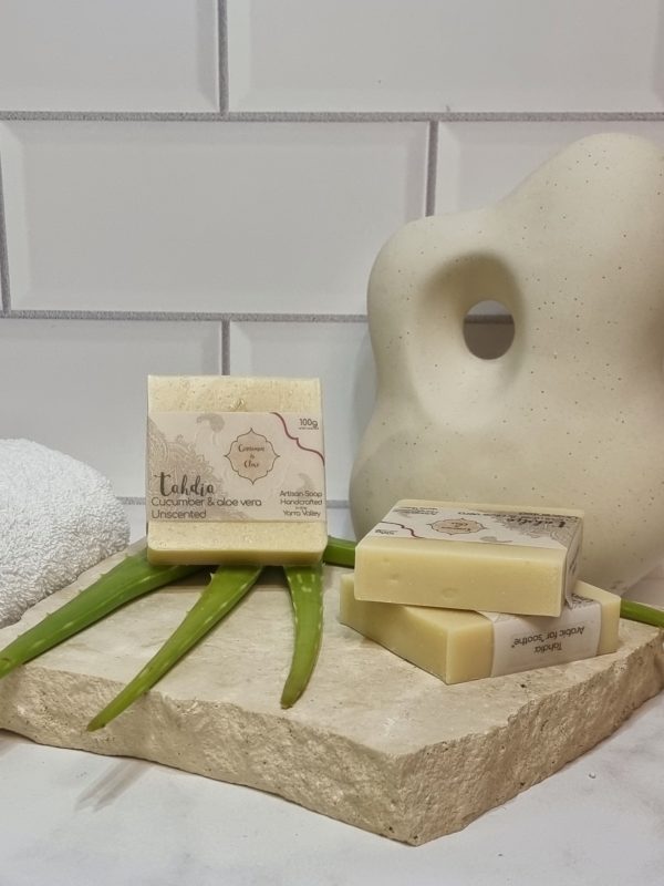 A tile background and stone bench with a piece of terrazzo. Three bars of creamy coloured homemade soap are sitting on the terrazo, with fresh aloe vera to decorate. Also in the background are a small white towel and a curvy decorative statue