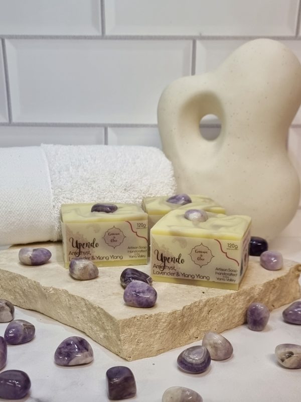 A tile background with a stone benchtop and a piece of terrazzo. Three bars of cream and purple swirled square soaps with a purple crystal in the top of each are sitting on the terrazzo with more purple crystals scattered around them. Also in the background are a small white towel and a curvy decorative statue