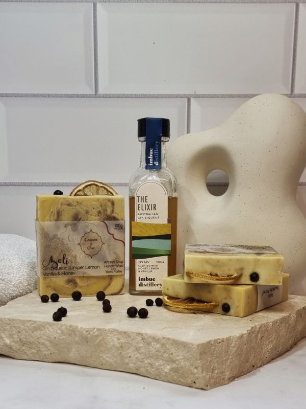 A tile background and stone bench with a piece of terrazzo. Three bars of yellow, brown and white swirled homemade soap with a lemon slice and juniper berry on top are sitting on the terrazzo with juniper berries scattered around them, and a bottle of Imbue Distillery The Elixr Liqueur. Also in the background are a small white towel and a curvy decorative statue