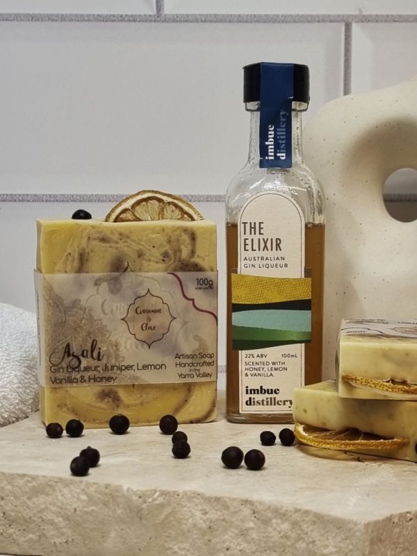 A tile background and stone bench with a piece of terrazzo. Three bars of yellow, brown and white swirled homemade soap with a lemon slice and juniper berry on top are sitting on the terrazzo with juniper berries scattered around them, and a bottle of Imbue Distillery The Elixr Liqueur. Also in the background are a small white towel and a curvy decorative statue