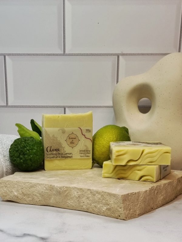 A tile background and stone bench with a piece of terrazzo. Three bars of creamy coloured homemade soap with a textured top are sitting on the terrazzo with a lemon and kaffir lime behind them. Also in the background are a small white towel and a curvy decorative statue