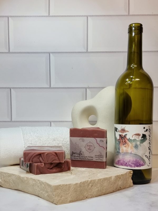 A tile background and stone bench with a piece of terrazzo. Three bars of red wine coloured homemade soap with a textured top are sitting on the terrazzo with a bottle of Payten and Jones Sangiovese behind them. Also in the background are a small white towel and a curvy decorative statue