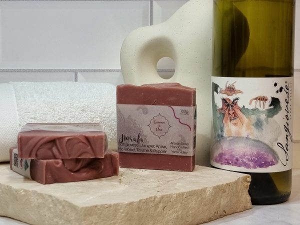 A tile background and stone bench with a piece of terrazzo. Three bars of red wine coloured homemade soap with a textured top are sitting on the terrazzo with a bottle of Payten and Jones Sangiovese behind them. Also in the background are a small white towel and a curvy decorative statue
