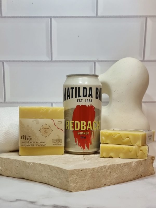 A tile background and stone bench with a piece of terrazzo. Three bars of cream coloured homemade soap with a textured top are sitting on the terrazzo with a can of Matilda Bay Redback beer behind them. Also in the background are a small white towel and a curvy decorative statue