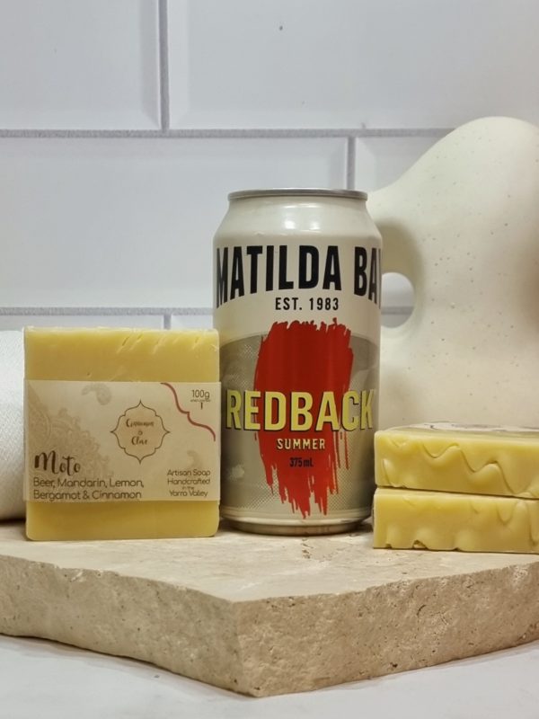 A tile background and stone bench with a piece of terrazzo. Three bars of cream coloured homemade soap with a textured top are sitting on the terrazzo with a can of Matilda Bay Redback beer behind them. Also in the background are a small white towel and a curvy decorative statue