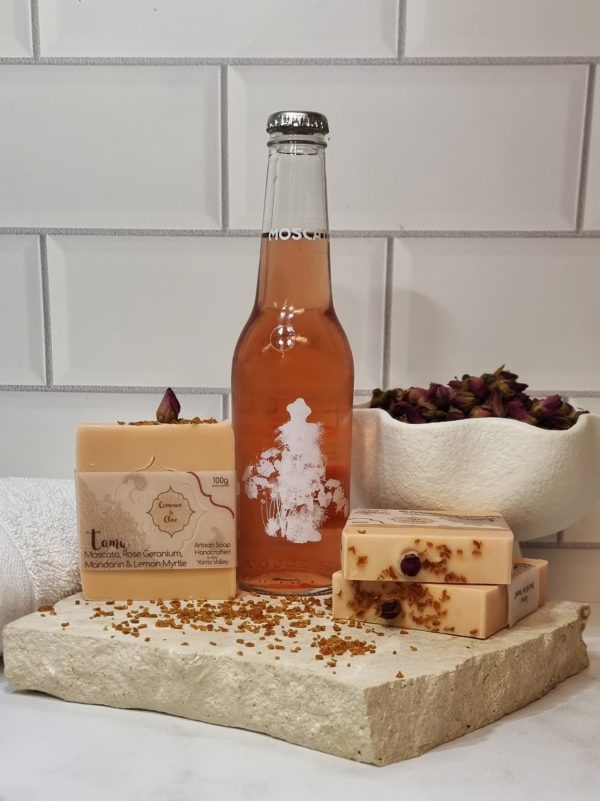 A tile background and stone bench with a piece of terrazzo. Three bars of pink homemade soap with a red rose bud and sprinkle of orange peel on top are sitting on the terrazzo with orange peel scattered around them, and a bottle of Innocent Bystander Moscato. Also in the background are a small white towel and a white fluted bowl filled with red rose buds