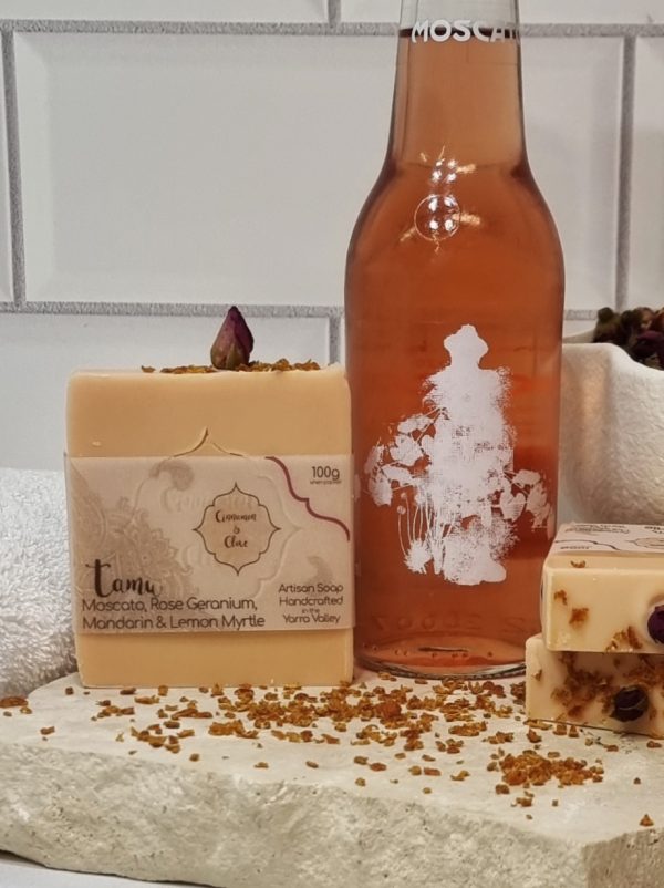 A tile background and stone bench with a piece of terrazzo. Three bars of pink homemade soap with a red rose bud and sprinkle of orange peel on top are sitting on the terrazzo with orange peel scattered around them, and a bottle of Innocent Bystander Moscato. Also in the background are a small white towel and a white fluted bowl filled with red rose buds