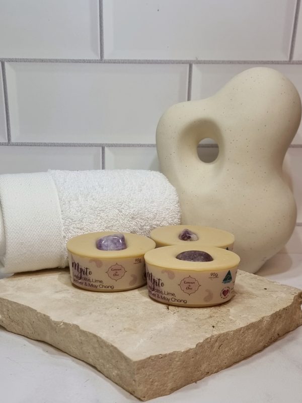 A tile background with a piece of terrazzo. Three bars of pale purple coloured oval shaped soaps with a purple crystal in top are sitting on the terrazzo. Also in the background are a small white towel and a curvy decorative statue