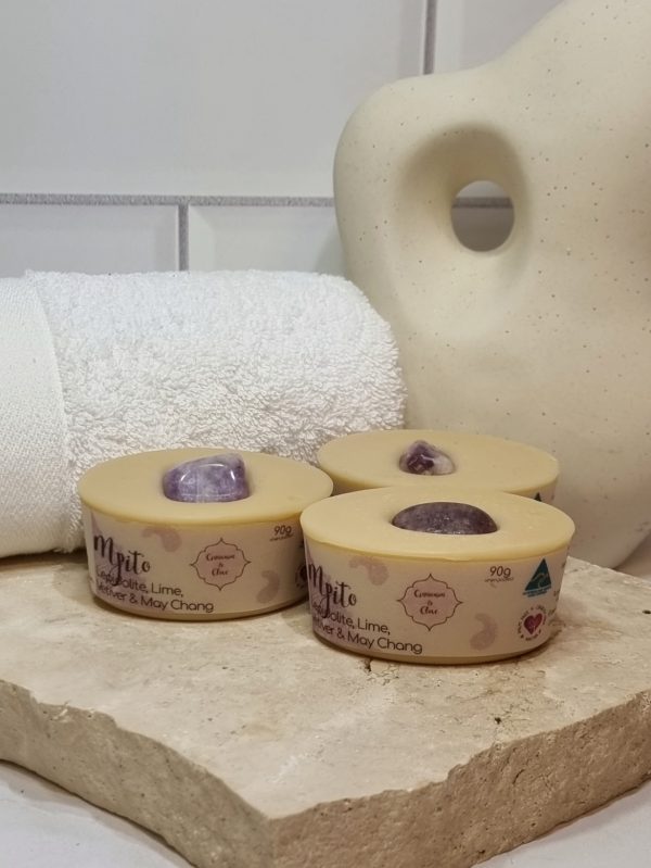 A tile background with a piece of terrazzo. Three bars of pale purple coloured oval shaped soaps with a purple crystal in top are sitting on the terrazzo. Also in the background are a small white towel and a curvy decorative statue