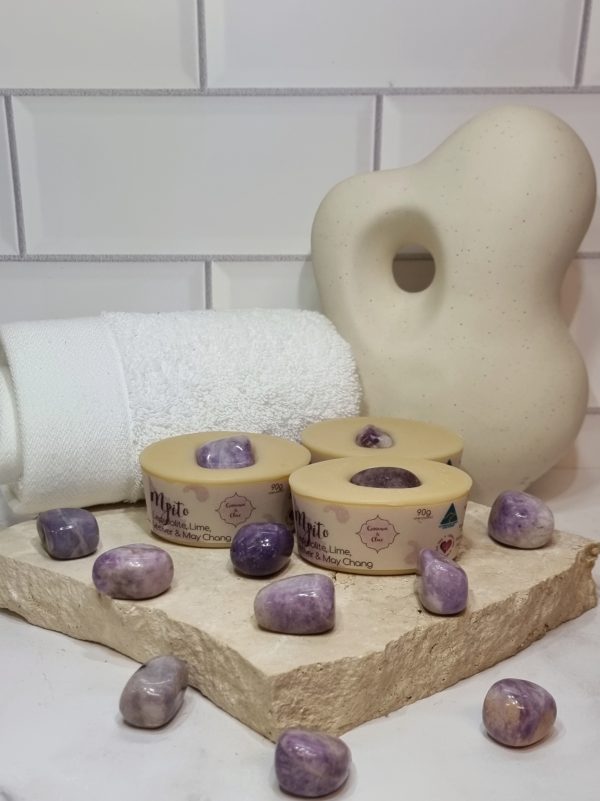 A tile background with a piece of terrazzo. Three bars of pale purple coloured oval shaped soaps with a purple crystal in top are sitting on the terrazzo with more purple crystals scattered around them. Also in the background are a small white towel and a curvy decorative statue